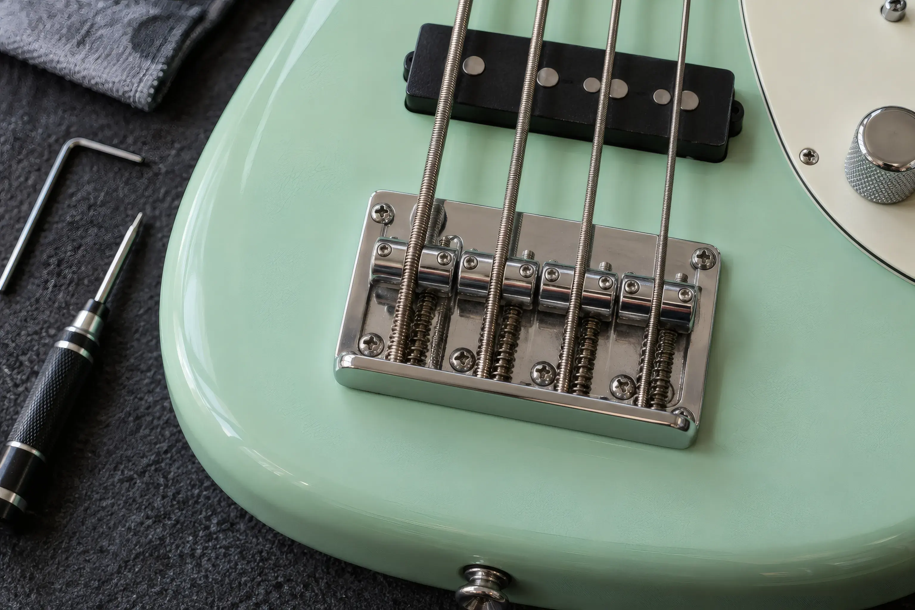 Close-up of four bass strings crossing individual chrome bridge saddles, with a hex key and screwdriver resting nearby for setup work